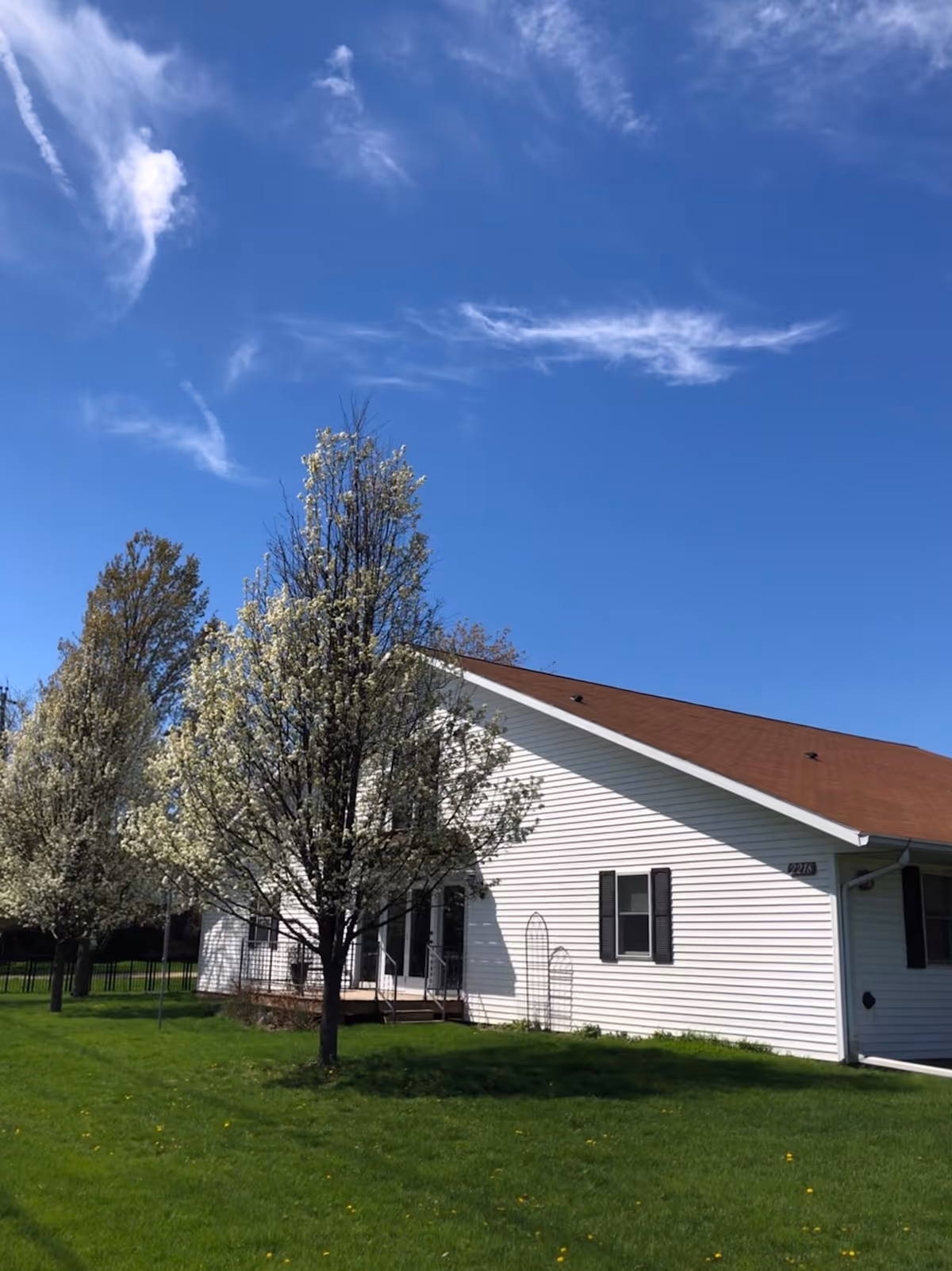 Exterior view of a single-story white building with a brown roof, surrounded by green grass and several blossoming trees under a blue sky with scattered clouds.