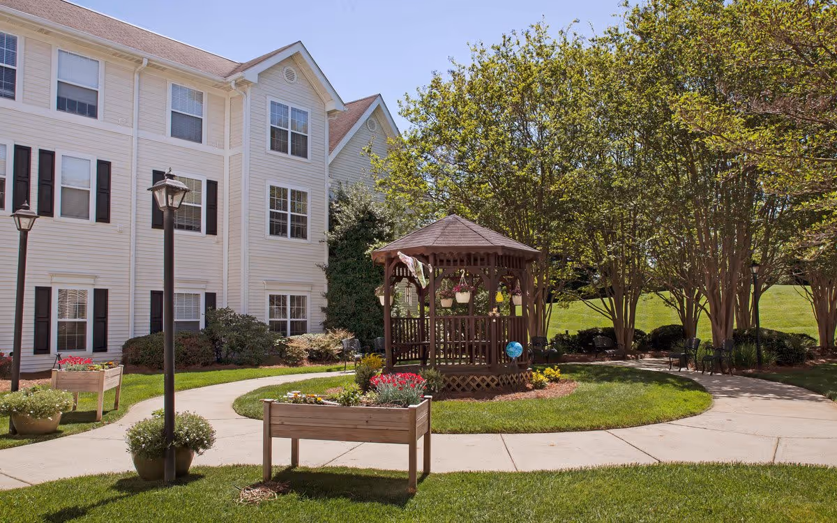 Courtyard with a wooden gazebo, winding sidewalks, planters and lawn in front of a multi-story senior living building.