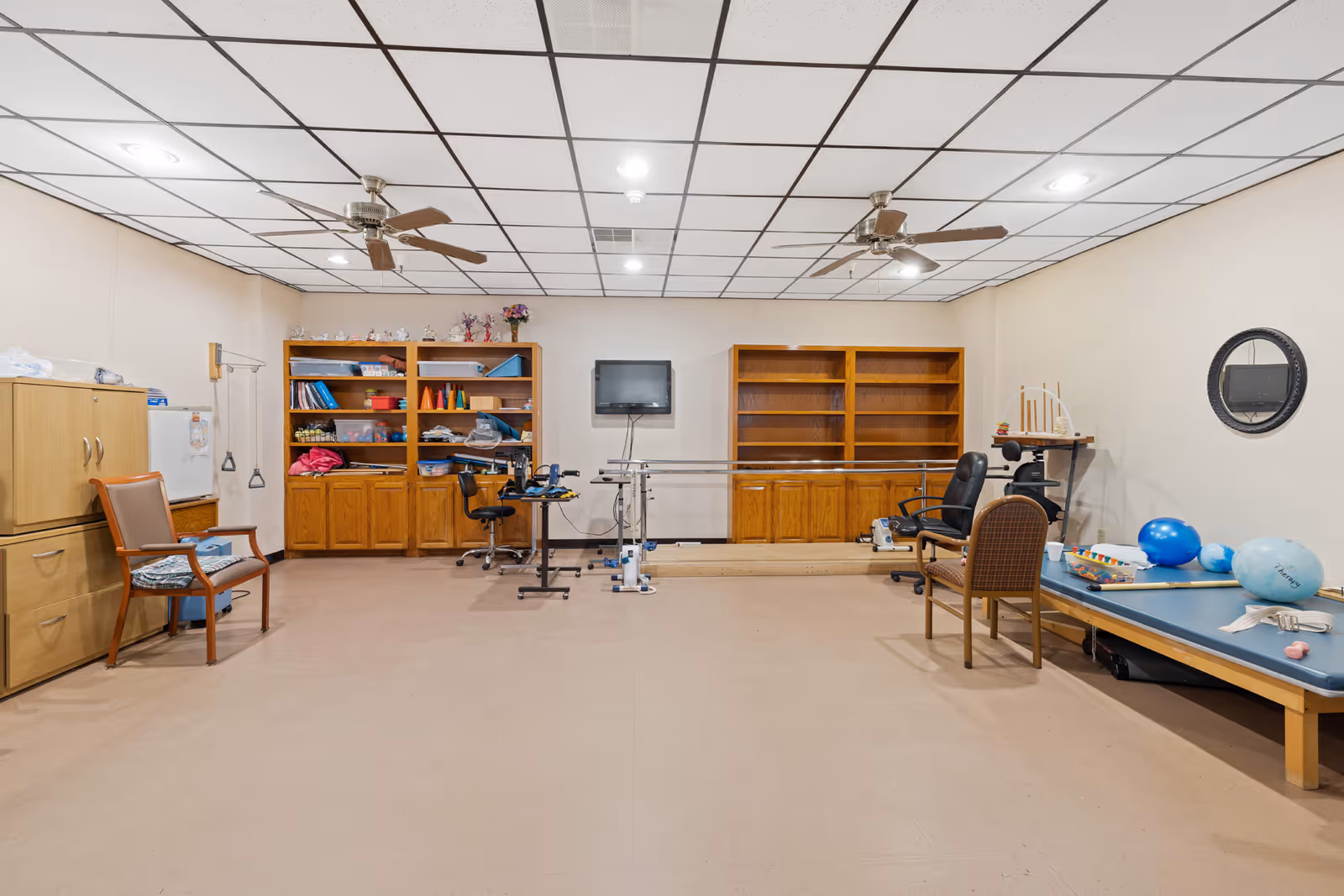 A spacious therapy or activity room with beige walls and a tiled ceiling featuring two ceiling fans. The room contains wooden cabinets and shelves filled with various supplies, a mounted TV, exercise equipment including parallel bars, chairs, and a therapy table with exercise balls and other therapy tools.