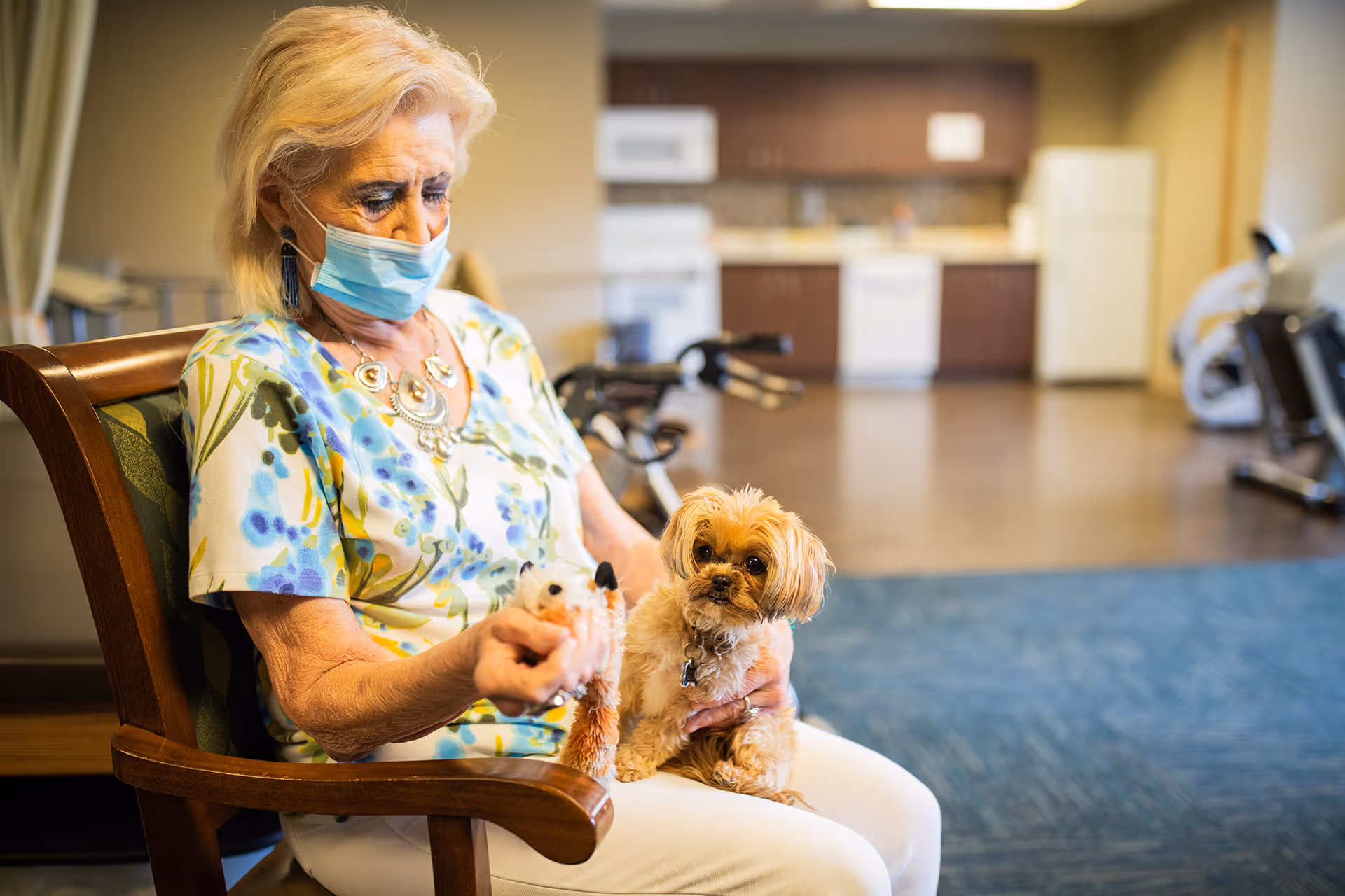 An elderly woman wearing a face mask sits on a wooden chair indoors, holding a small dog on her lap and a toy in her hand. The background shows a kitchen area with cabinets, a refrigerator, and exercise equipment.