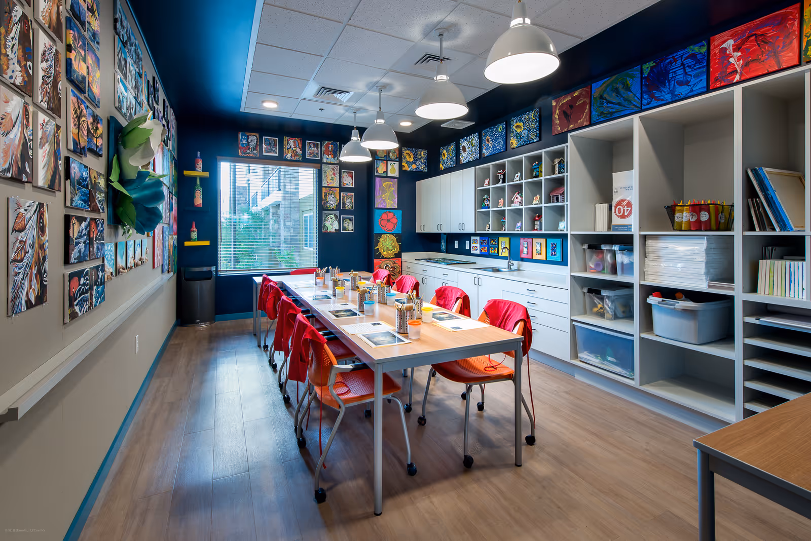 Bright, colorful art and craft room with a long table surrounded by red chairs, shelving of supplies, and artwork on the walls.