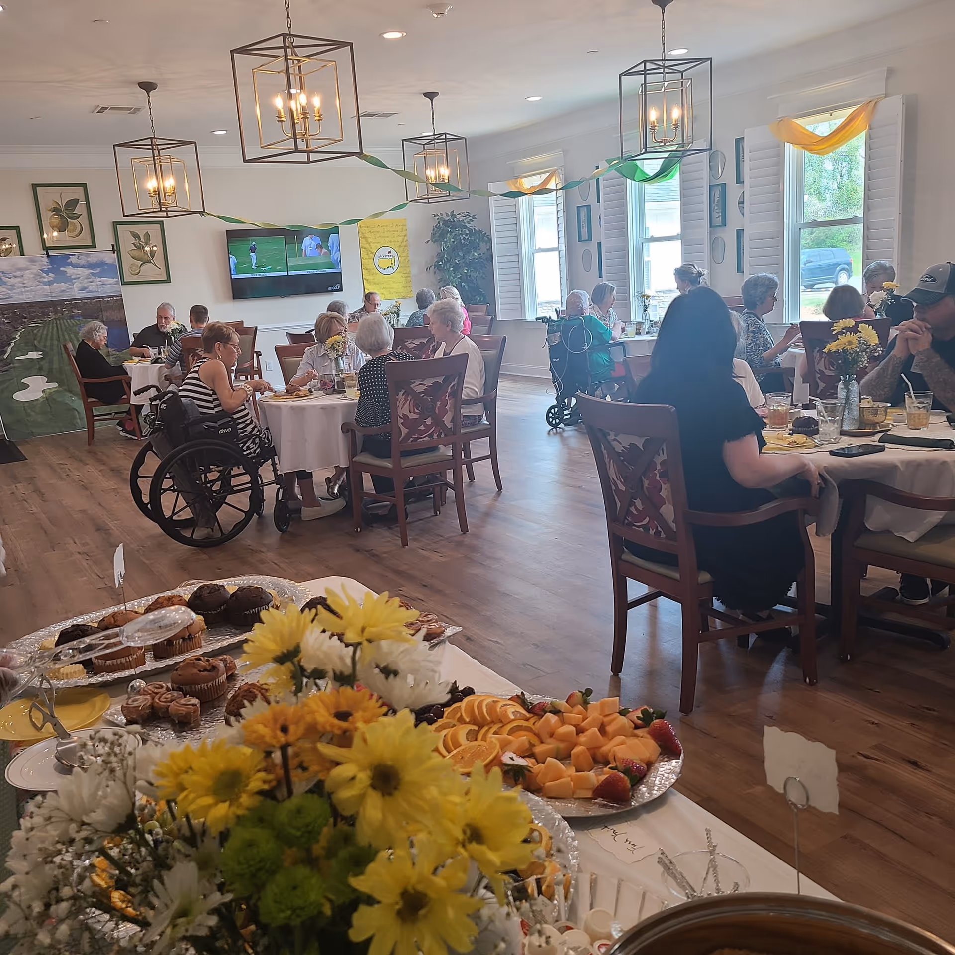 A dining room in a senior living facility with several elderly residents seated at round tables enjoying a meal. The room is decorated with hanging light fixtures and streamers. A table in the foreground displays a variety of muffins, pastries, and fresh fruit platters with yellow and white flowers. Large windows let in natural light, and a television is mounted on the wall showing a golf game.