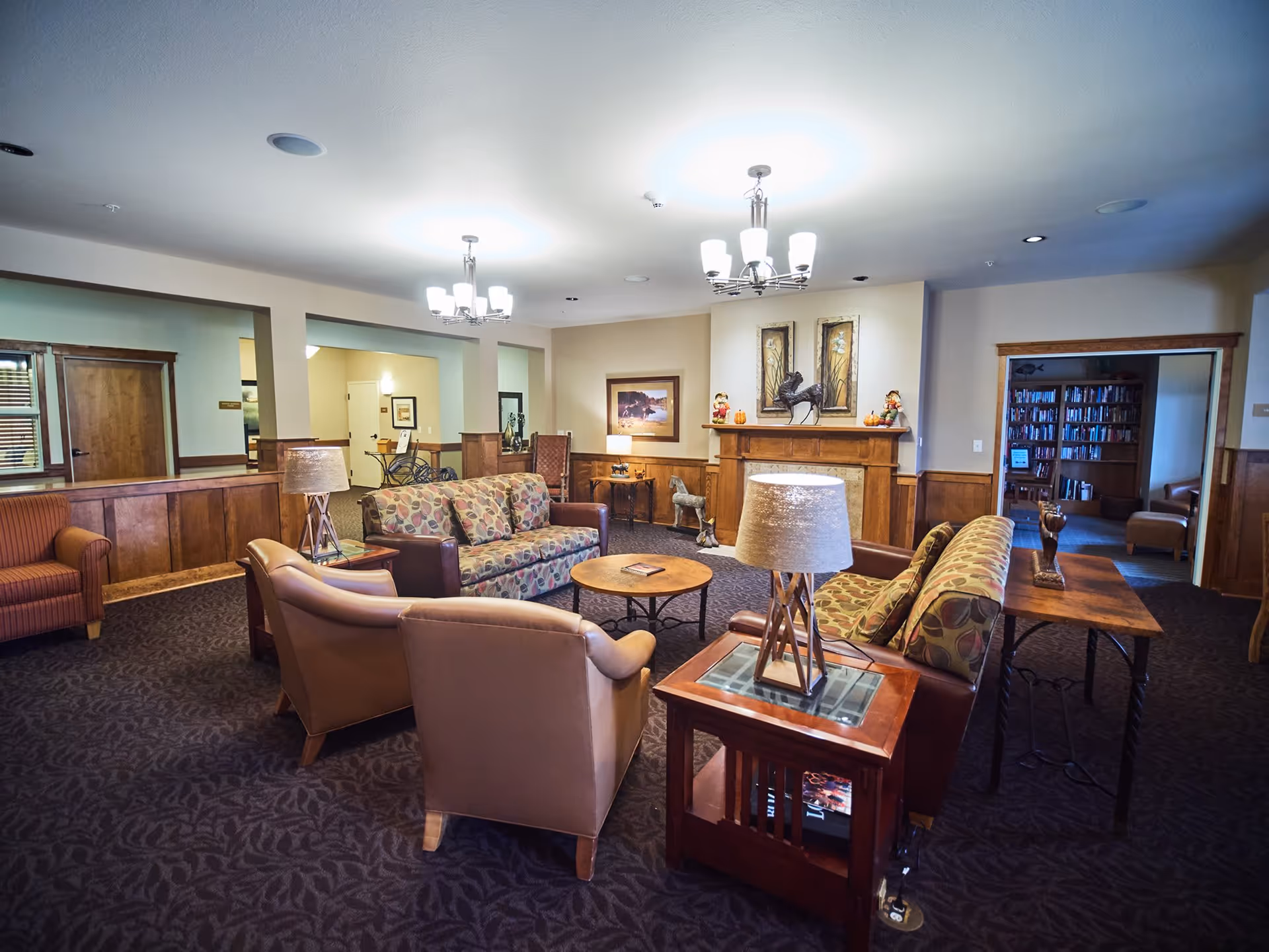 Spacious communal living room with sofas, armchairs, coffee tables, lamps, and a mantel/fireplace in a senior facility.