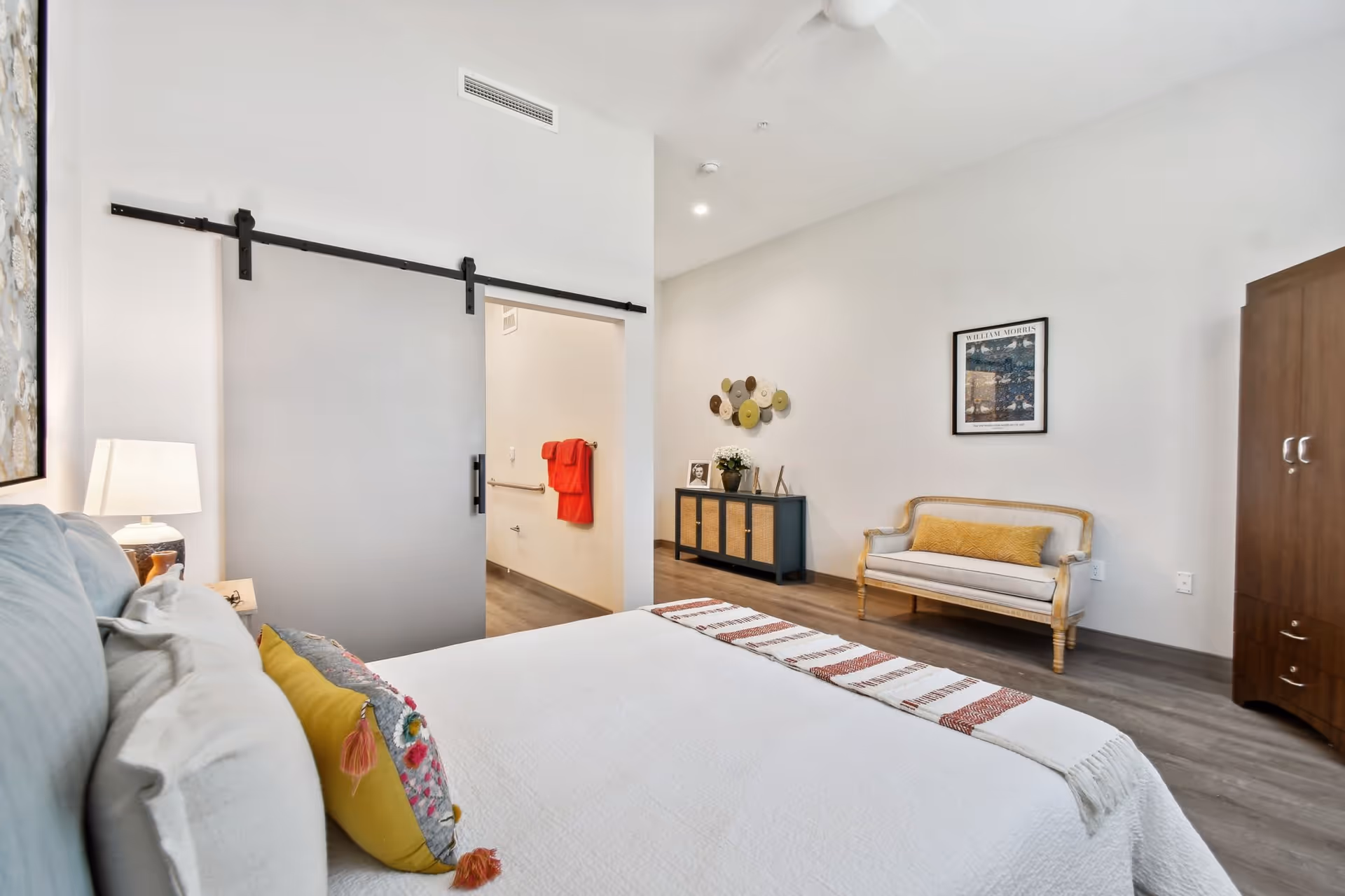 A bright and modern bedroom with a large bed featuring white bedding and colorful pillows. There is a sliding barn door leading to a bathroom with red towels visible. The room also includes a small sofa with a yellow cushion, a dark cabinet with decorative items, and a wooden wardrobe. The walls are white and the floor is a light wood finish.
