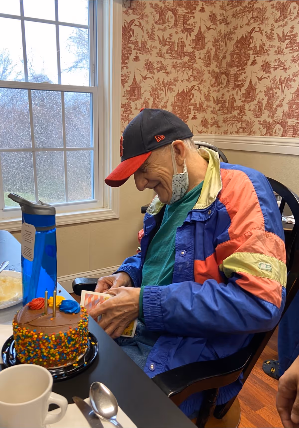 An elderly man wearing a colorful jacket and a baseball cap is sitting at a table near a window with a birthday cake decorated with colorful sprinkles and candles in front of him. He is looking down and smiling while holding a card. There is a blue water bottle and a white cup with a spoon on the table.