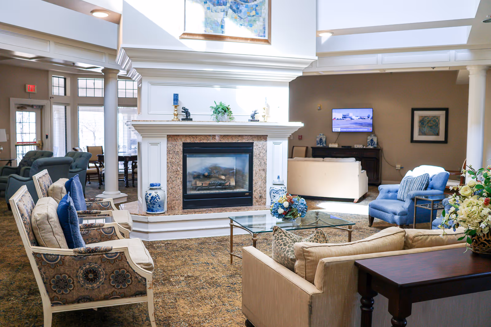 Bright senior living common lounge with a central fireplace, sofas, armchairs, glass coffee table, and a wall-mounted TV.