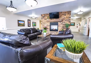 A cozy living room area featuring three dark leather sofas arranged around a glass coffee table with a plant centerpiece. A stone fireplace with a mounted flat-screen TV above it is centered on the far wall. The room is well-lit with ceiling lights and decorated with framed landscape paintings on the walls. A side table with a potted plant and books is visible in the foreground.