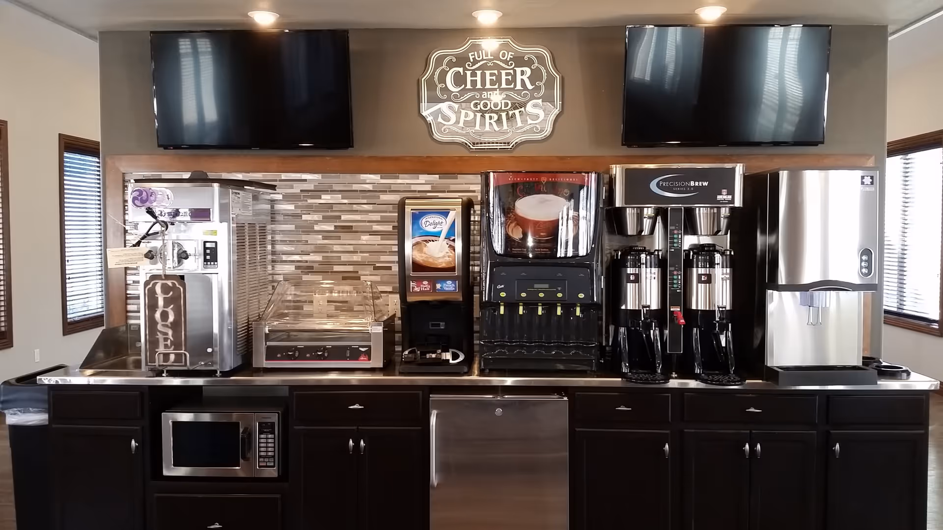 A beverage station with various drink machines including a frozen drink machine with a 'CLOSED' sign, a creamer dispenser, a hot beverage dispenser, two coffee urns, and an ice machine. Above the station are two large flat-screen TVs and a decorative sign that reads 'Full of Cheer and Good Spirits'. The station has dark cabinetry and a stainless steel countertop, with windows on either side letting in natural light.