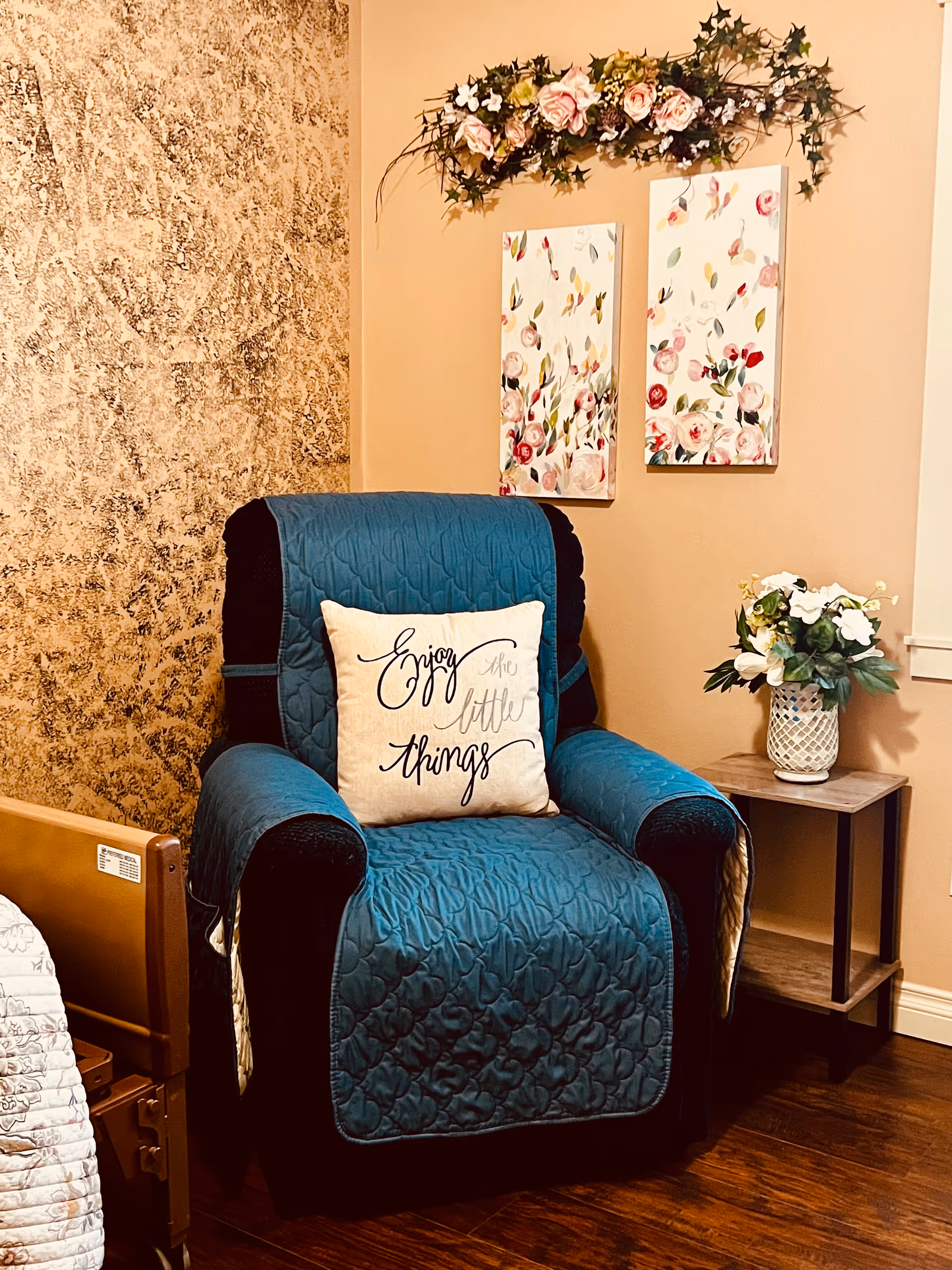 A cozy corner of a room featuring a dark armchair covered with a teal quilted cover and a decorative pillow that says 'Enjoy the little things'. Behind the chair, there are two floral paintings on the wall with a floral garland above them. To the right of the chair is a small wooden side table with a white vase holding white and green flowers. The floor is wooden, and part of a bed frame is visible on the left side.