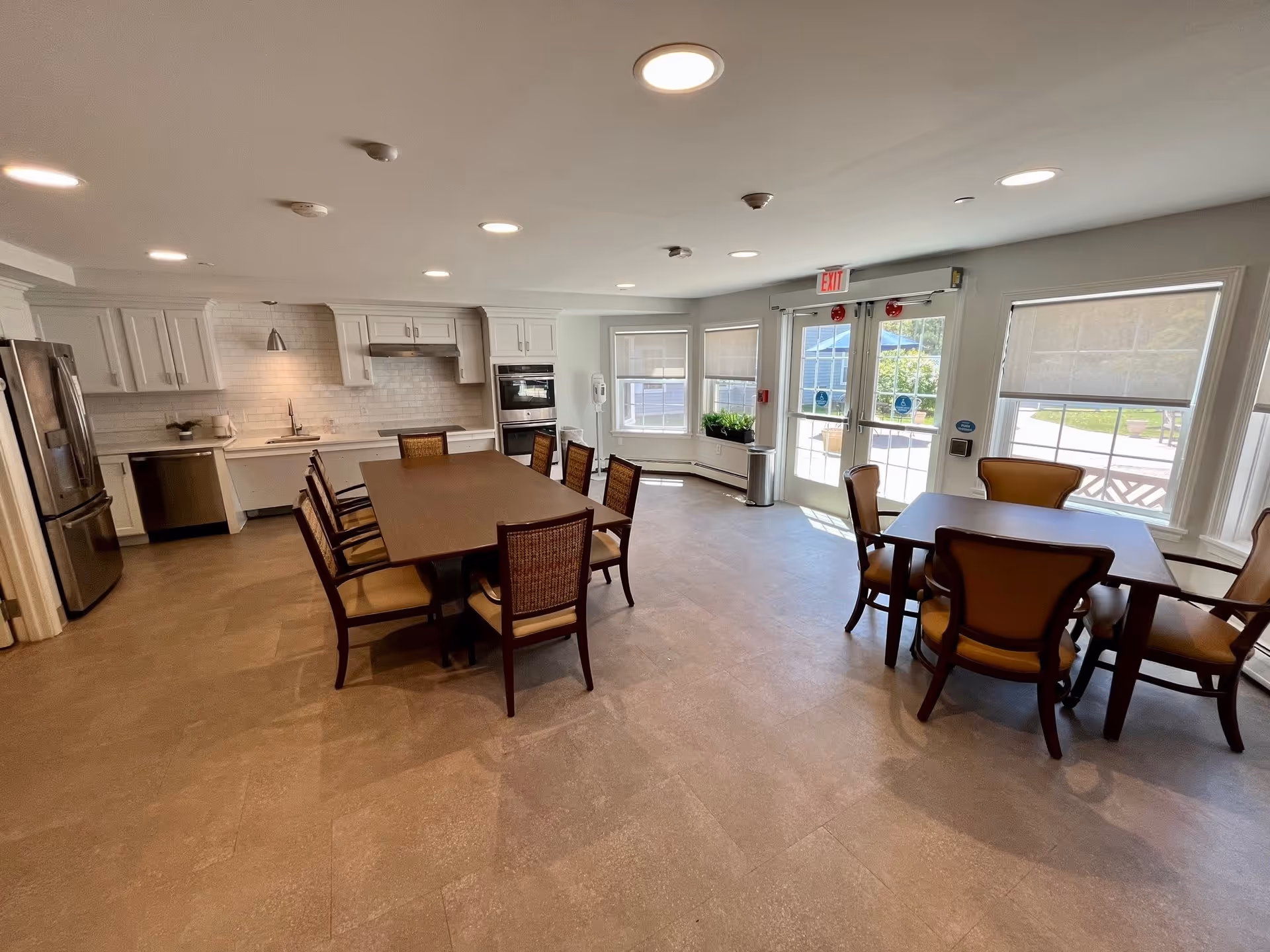 Bright communal dining area with a long table and chairs, a full kitchen along one wall, and large windows and doors letting in natural light.