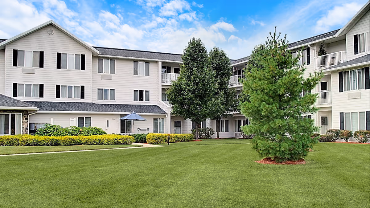 Exterior view of a three-story senior living facility building with white siding and black shutters, surrounded by a well-maintained green lawn, bushes, and several trees under a partly cloudy blue sky.