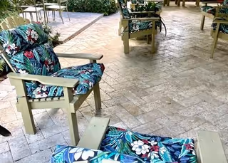 Outdoor shaded patio with cushioned wooden chairs featuring tropical floral cushions on a brick-tile floor.