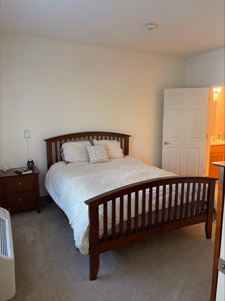 A bedroom with a wooden bed frame and white bedding, including three pillows. There is a wooden nightstand with an alarm clock and a book on it. The room has beige carpet and light-colored walls. An open door reveals a bathroom with a sink and mirror.
