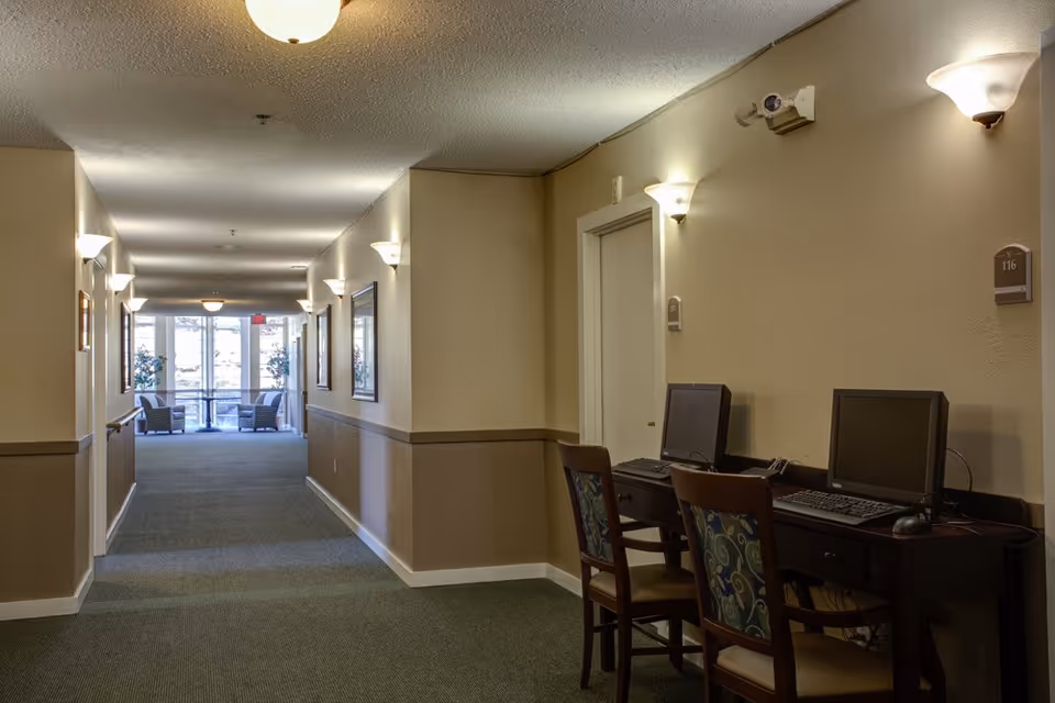 Carpeted interior hallway of an assisted living facility with wall sconces, two computer workstations on the right, and seating visible at the far end.