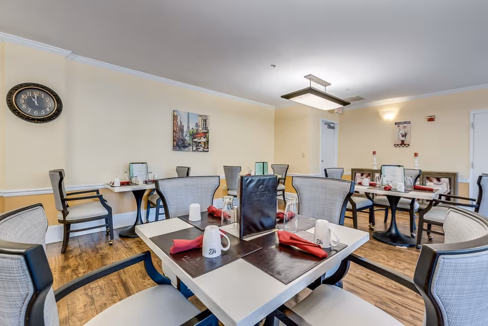 A dining room in a senior living facility with several tables set with placemats, red napkins, white mugs, and glasses. The room has light yellow walls, wooden flooring, and a wall clock. There are framed pictures on the walls and modern chairs around the tables.