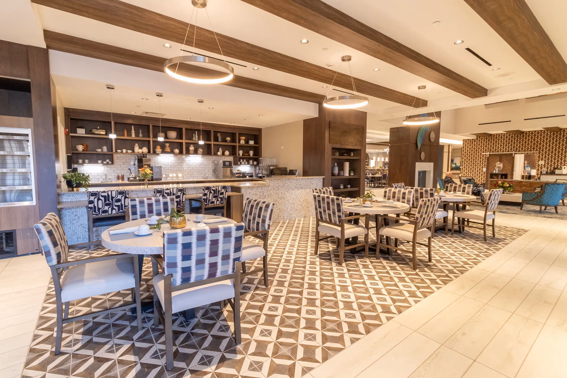 Bright communal dining area with round tables, patterned tile floor, upholstered chairs, and a countertop service area with open shelving.