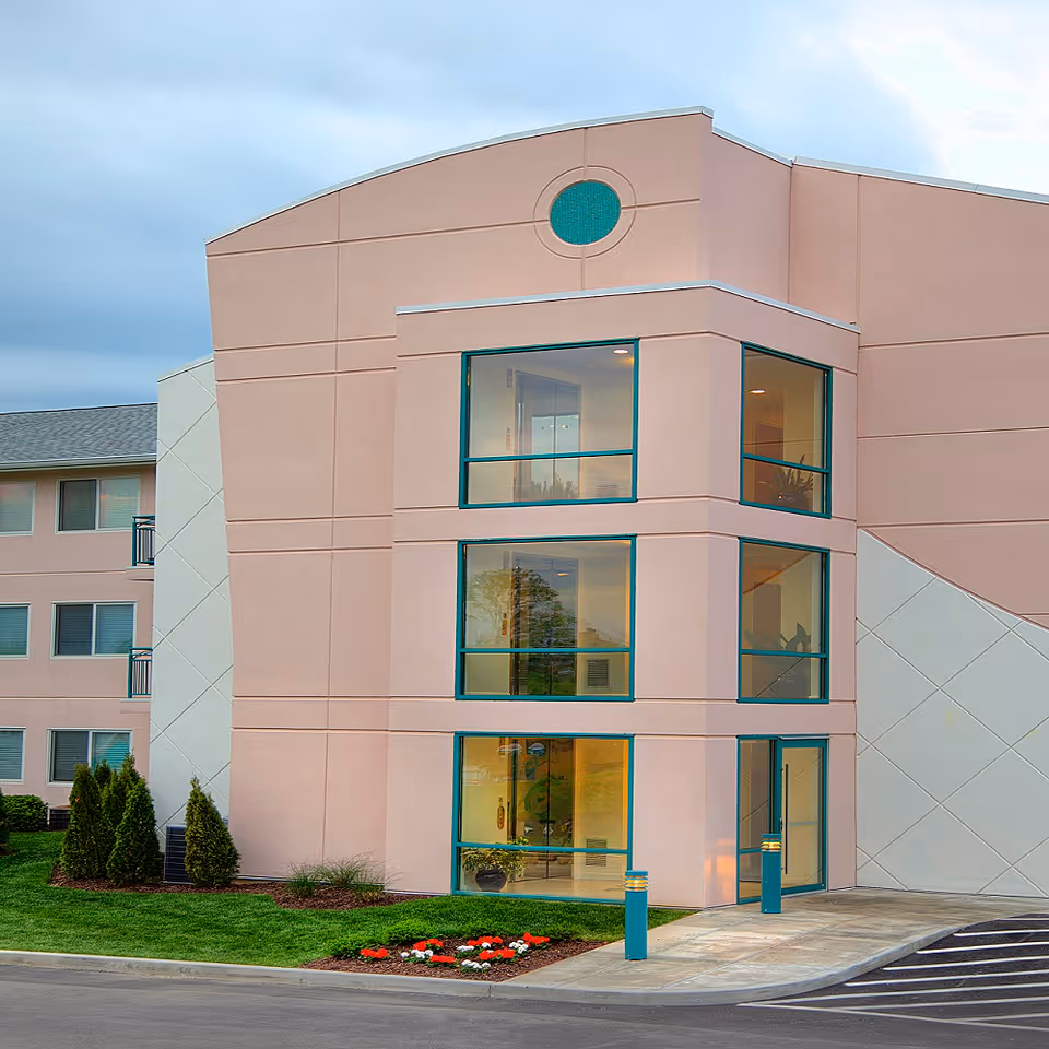 Modern pale-pink multi-story building front with large glass windows, an entrance and a small landscaped flower bed.