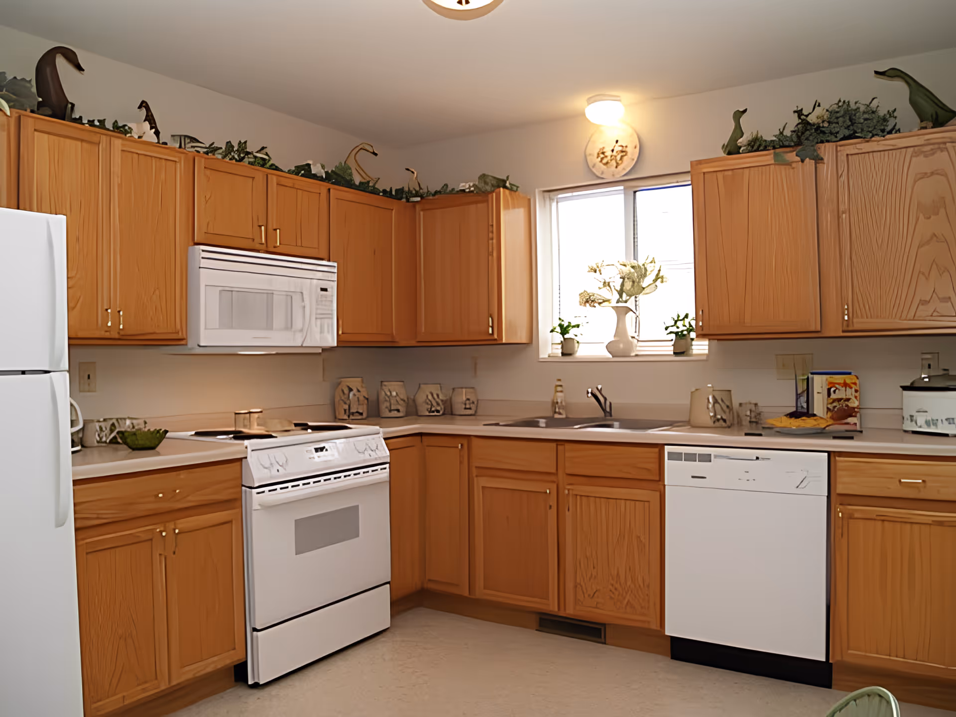 Sunny kitchen with light wood cabinets, white appliances including stove, microwave, refrigerator and dishwasher, and a sink beneath a window with potted plants.