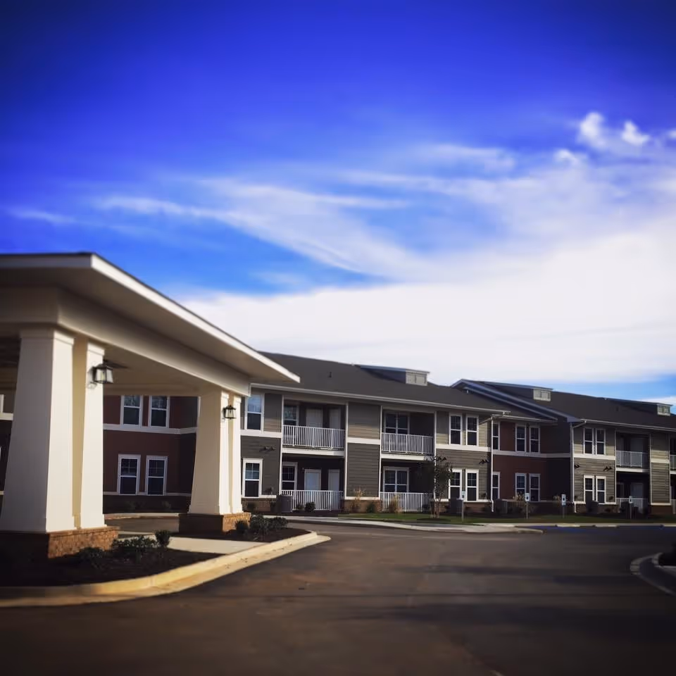 Exterior view of a modern senior living facility building with multiple windows and balconies under a blue sky with some clouds. The foreground shows a covered entrance with white pillars and a paved driveway.