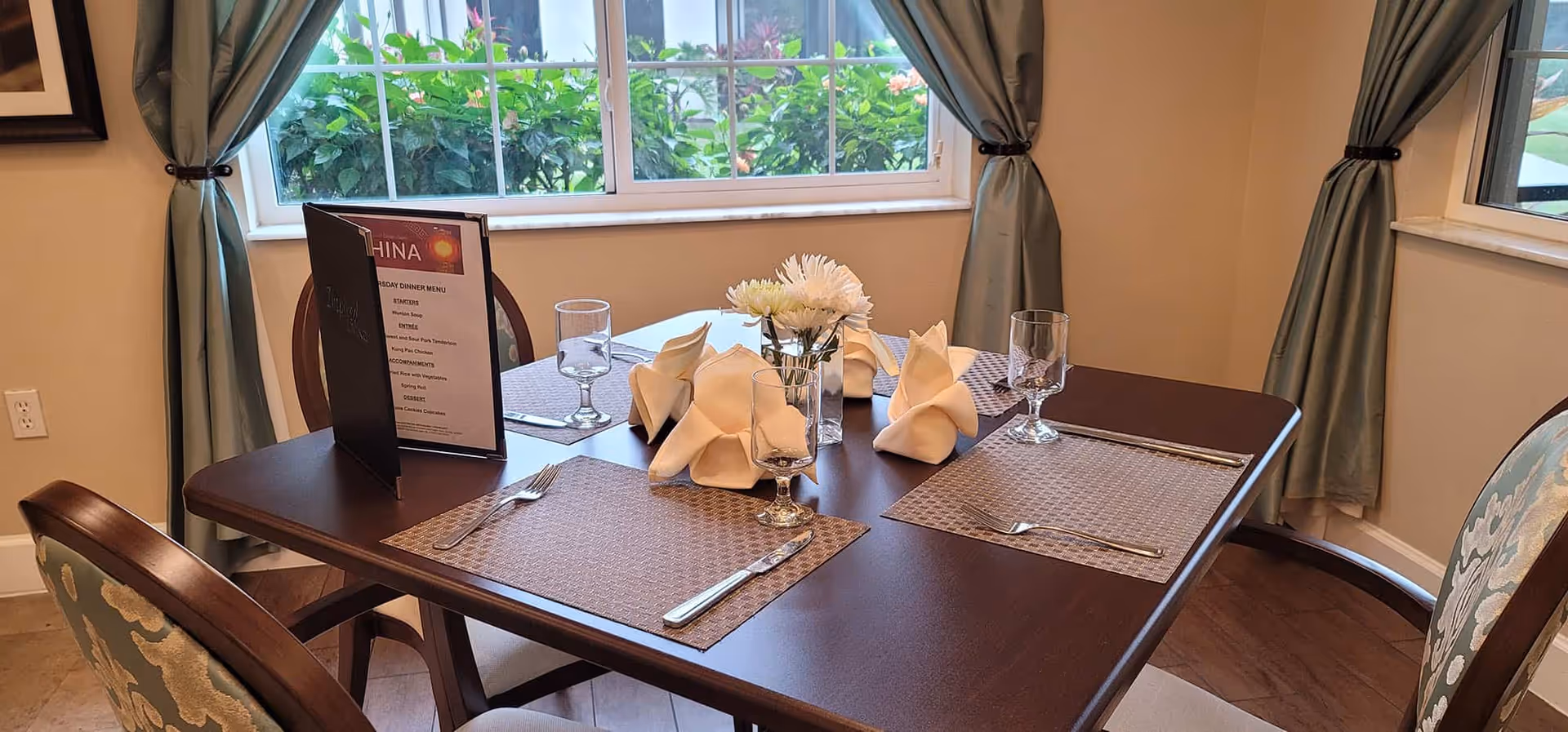 A dining table set for four with folded napkins, placemats, silverware, and glasses. A small vase with white flowers is in the center. Behind the table is a window with green plants outside and light blue curtains tied back on either side.