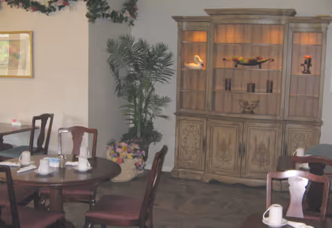 Interior view of a dining area with round wooden tables and chairs. Each table is set with white cups and napkins. There is a large wooden cabinet with decorative items and lighting in the background, along with a tall potted plant and floral arrangement.