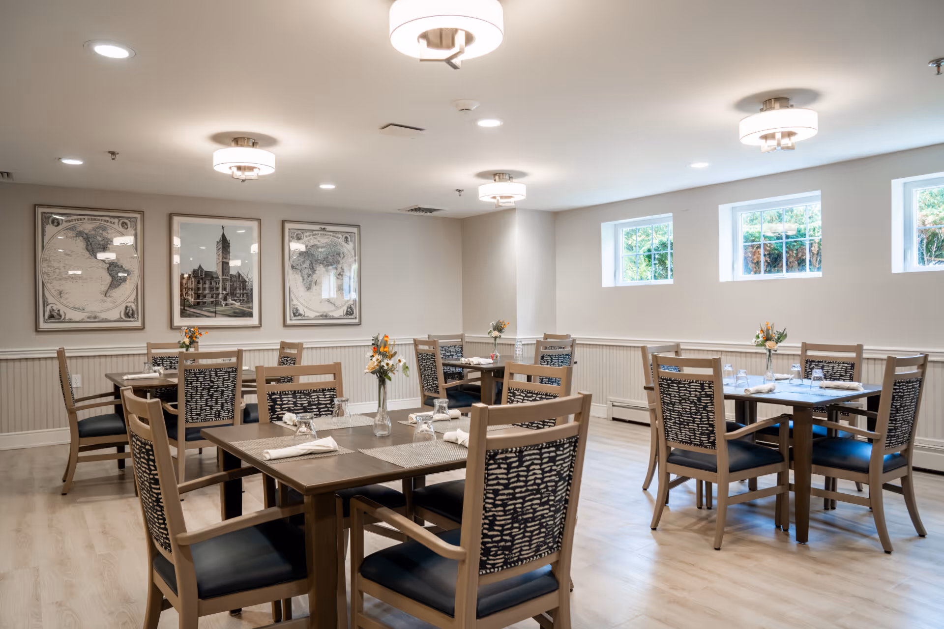 A bright dining room with several wooden tables and chairs arranged neatly. Each table has placemats, napkins, glasses, and small vases with flowers. The room has light-colored walls with three framed pictures hanging on one wall and three small windows letting in natural light. The floor is light wood, and modern ceiling lights illuminate the space.