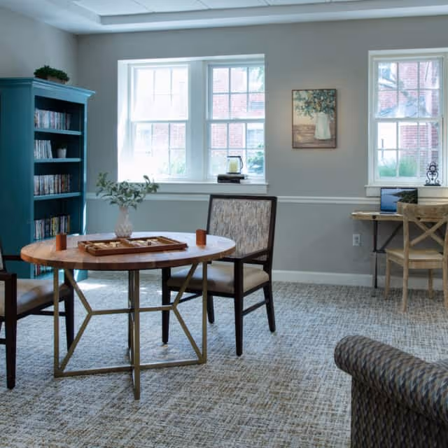 Bright communal sitting area with a round wooden table and chairs, a bookshelf, windows, and a small desk.