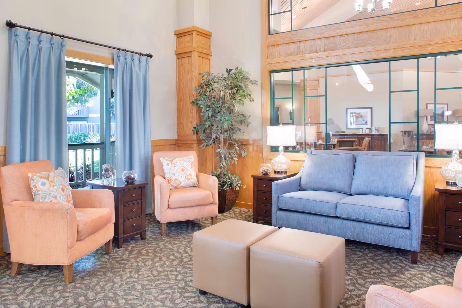 A cozy living room area with a blue sofa, two peach armchairs with patterned cushions, two beige ottomans, two dark wooden side tables with glass lamps, a large potted plant, and a window with blue curtains showing an outdoor view.