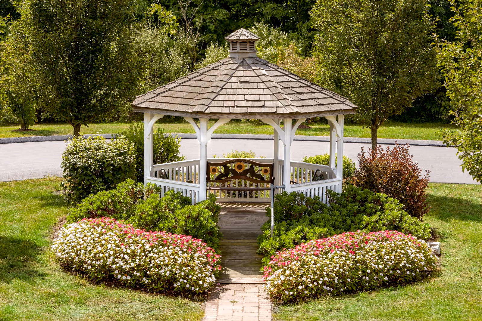 A white wooden gazebo with a shingled roof surrounded by green bushes and colorful flowers, situated on a grassy area with trees and a paved road in the background.