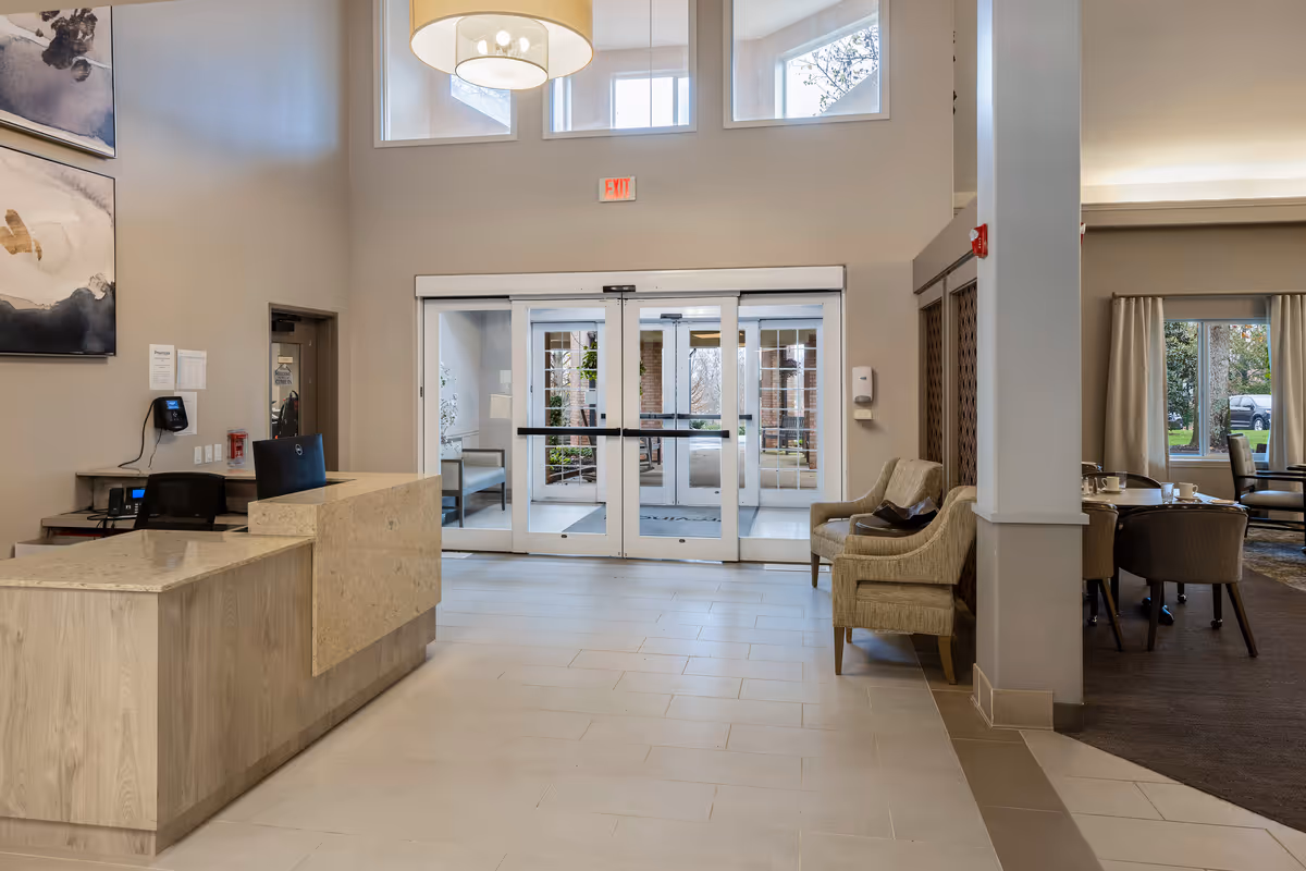 Interior view of a senior living facility lobby with a reception desk on the left, two armchairs on the right, and glass double doors leading outside. The space has high ceilings with windows above the doors, modern light fixtures, and neutral-colored walls and flooring.