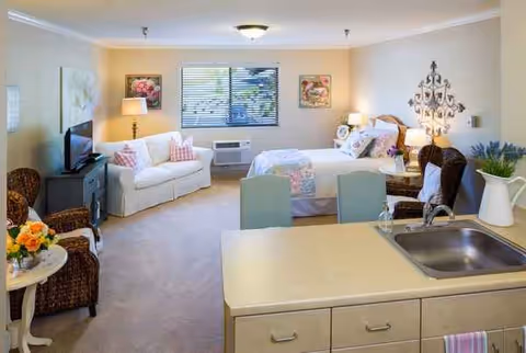 A bright and cozy assisted living studio apartment featuring a bed with floral bedding, a white loveseat with pillows, a TV on a stand, wicker chairs, a small table with flowers, and a kitchen counter with a sink in the foreground. The room has beige walls, a window with blinds, and decorative wall art.