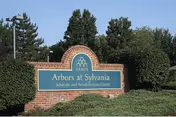 Brick entrance sign reading "Arbors at Sylvania Subacute and Rehabilitation Center" surrounded by shrubs and trees under a clear sky.