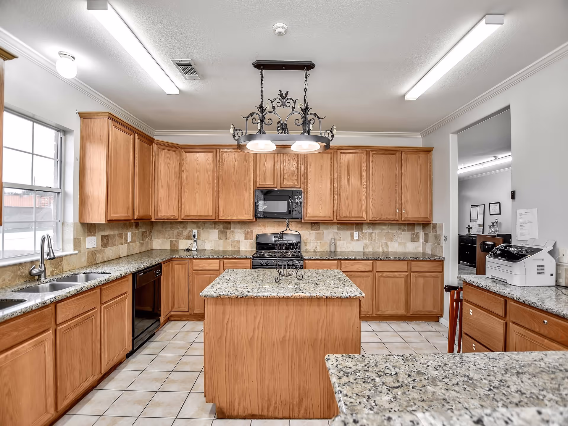 A spacious kitchen with wooden cabinets, granite countertops, and a central island. The kitchen features a double sink under a window, a black dishwasher, a black stove with a microwave above it, and tiled backsplash and floor. There is a decorative light fixture hanging above the island and fluorescent ceiling lights providing illumination.