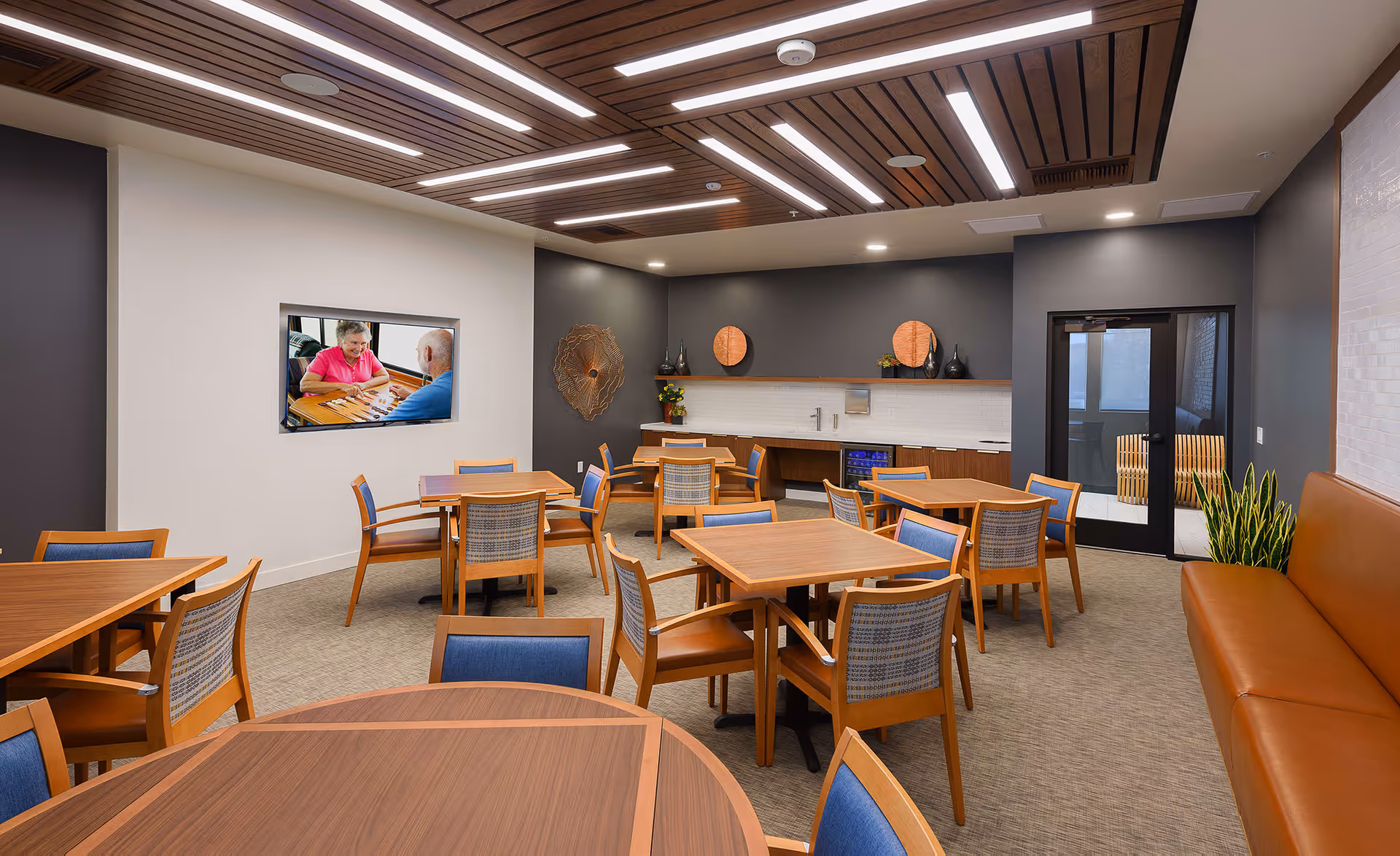 A modern dining room with multiple wooden tables and chairs with blue cushions. The room has a brown leather bench along one wall, a dark accent wall with decorative art pieces, and a countertop with a sink and small refrigerator. A flat-screen TV on a white wall displays an image of two elderly people playing a board game. The ceiling features wooden slats with integrated lighting.