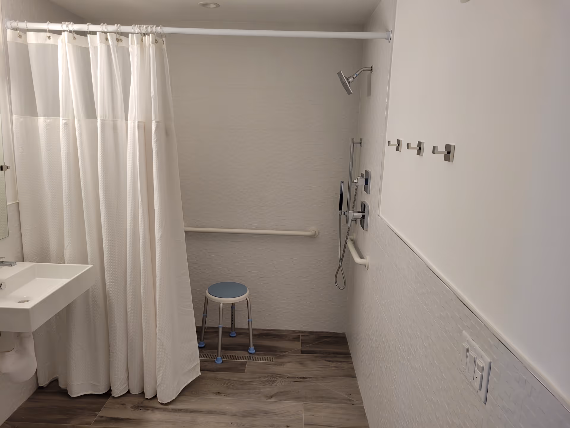 A clean, accessible bathroom with a white shower curtain, a small blue and white shower stool, a wall-mounted shower head, grab bars, a white sink, and wooden floor tiles.
