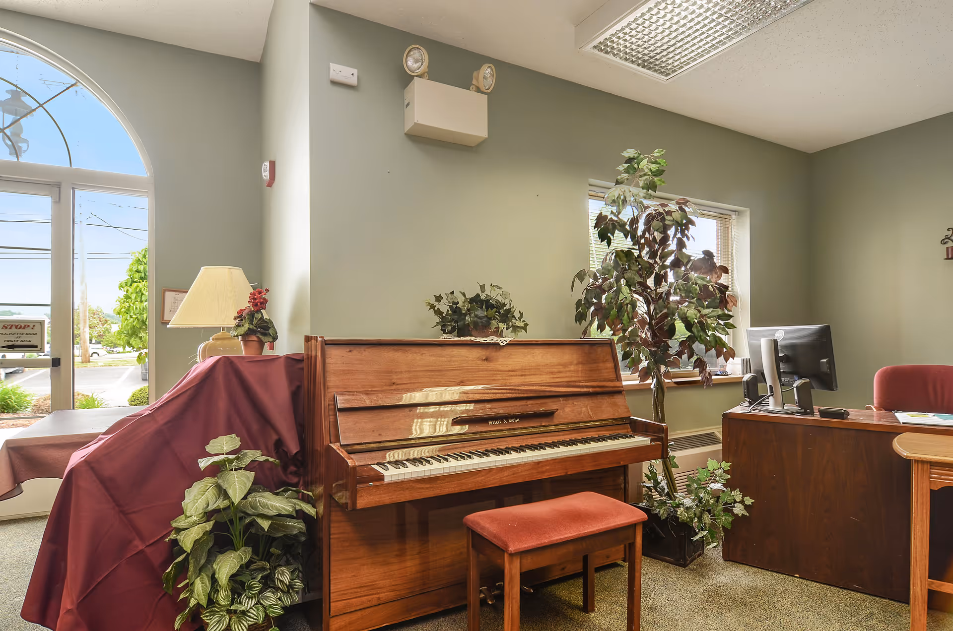 Interior view of a room with a wooden upright piano and matching bench in the center. To the left, a table covered with a burgundy cloth and a lamp with a beige shade sits near a large arched window and glass door showing an outdoor view. To the right, a wooden desk with a computer monitor and a red office chair is positioned near a window with blinds. Several green potted plants are placed around the room, which has light green walls and a ceiling with fluorescent lighting.