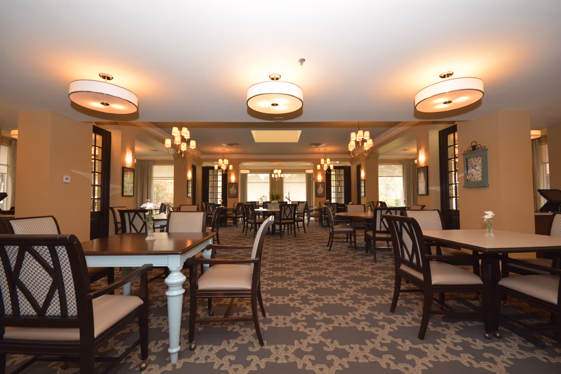 A spacious dining room with multiple tables and chairs arranged neatly. The room features patterned carpet flooring, beige walls, and large windows allowing natural light. Ceiling lights and wall sconces provide warm illumination. Small vases with flowers are placed on the tables.