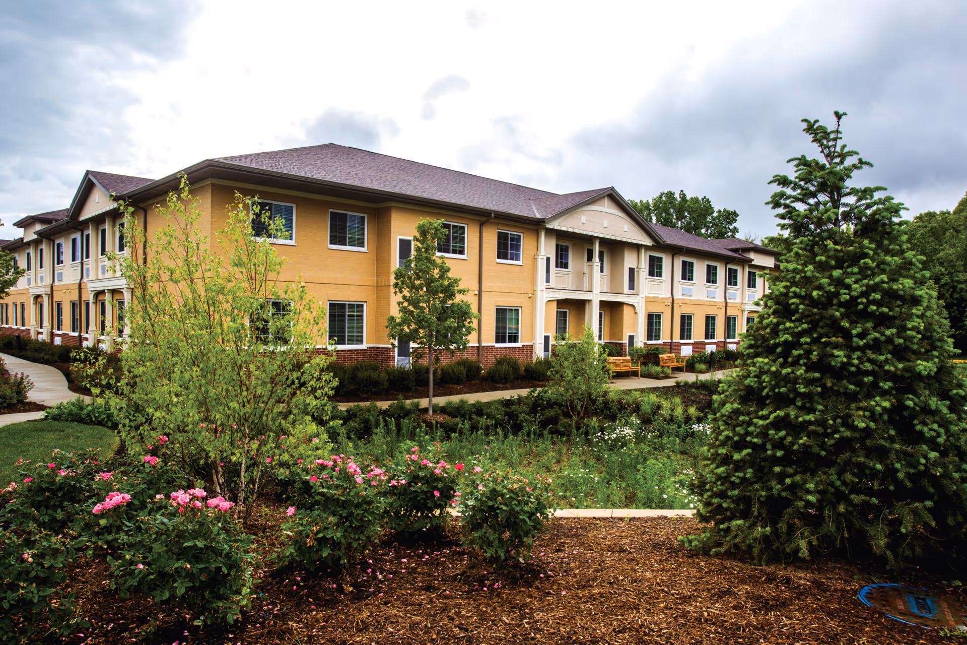 Exterior view of a two-story assisted living facility building with beige and brick facade, surrounded by landscaped gardens with green trees, bushes, and pink flowers under a cloudy sky.