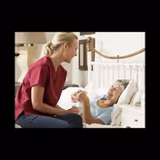 A caregiver in scrubs sits beside an elderly woman smiling in bed inside a bedroom.