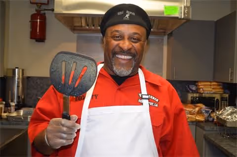 A smiling man wearing a black chef hat and a red shirt with a white apron is holding a large spatula in a kitchen setting with various kitchen appliances and food items in the background.