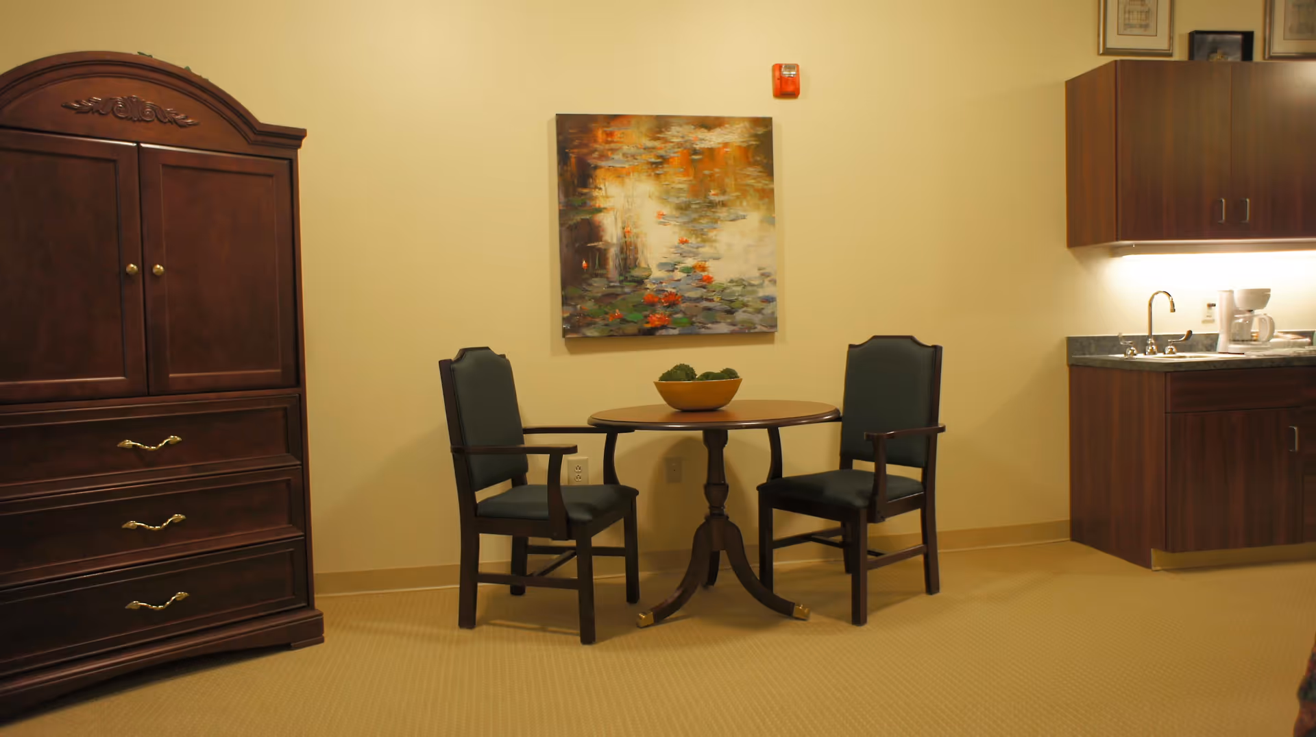 A small sitting area with a round wooden table and two dark wooden chairs with green cushions. On the table is a decorative bowl with green moss balls. Behind the table is a wall with a colorful abstract painting. To the left is a large dark wooden armoire with drawers, and to the right is a kitchenette area with a sink, coffee maker, and wooden cabinets.