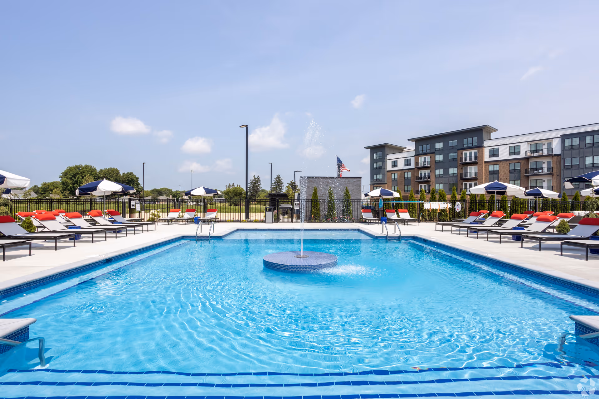Outdoor swimming pool with a central fountain, rows of lounge chairs and umbrellas, and a multi-story building in the background.
