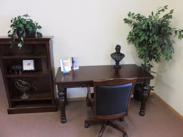 A small office or study area with a dark wooden desk and a black leather office chair. On the desk are two small framed pictures and a bust sculpture. To the left of the desk is a matching dark wooden bookshelf with decorative items and a potted plant on top. To the right of the desk is a tall green leafy plant. The walls are light-colored and the floor is carpeted.