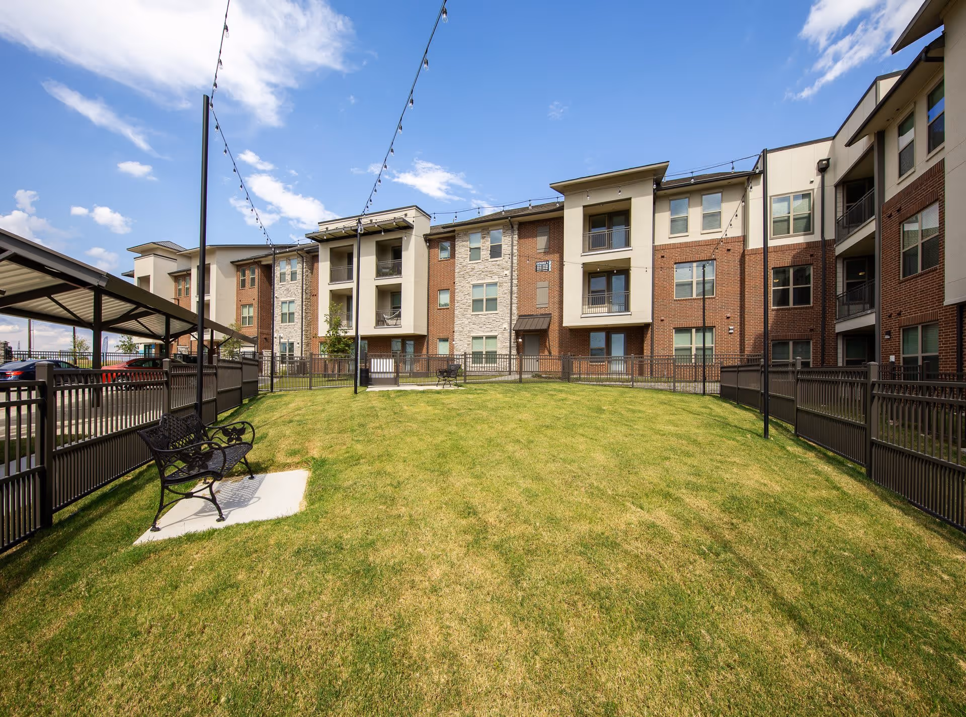 Grassy fenced courtyard with benches and string lights in front of a multi-story apartment building.