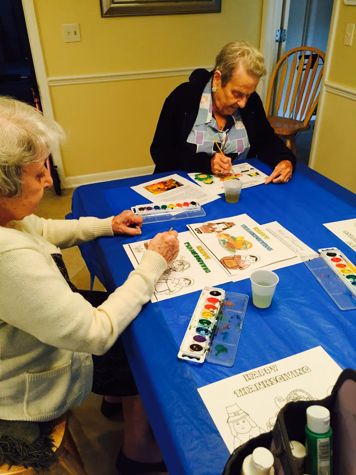 Two elderly women sitting at a table covered with a blue tablecloth, painting Thanksgiving-themed coloring pages using watercolor paints. The room has light yellow walls and wooden chairs.