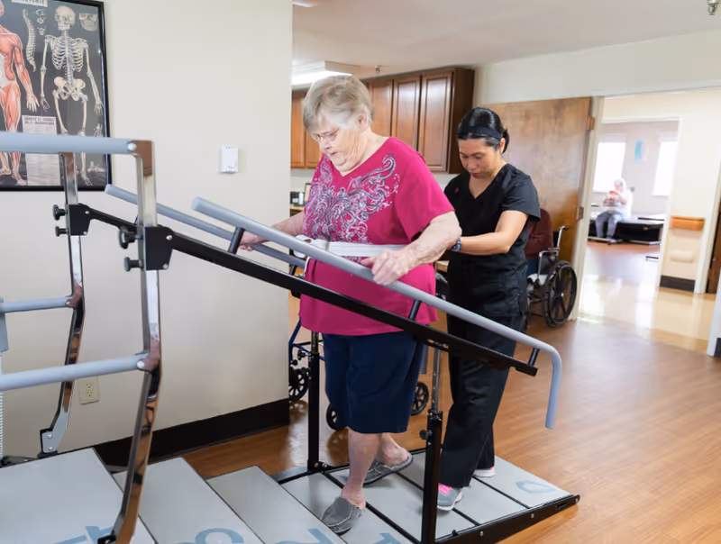 An elderly woman wearing a pink shirt and navy shorts is walking up a set of rehabilitation stairs with handrails, assisted by a healthcare worker dressed in black scrubs. The setting appears to be a healthcare or senior living facility with wooden floors and medical posters on the wall. In the background, another elderly person is seated near a window.