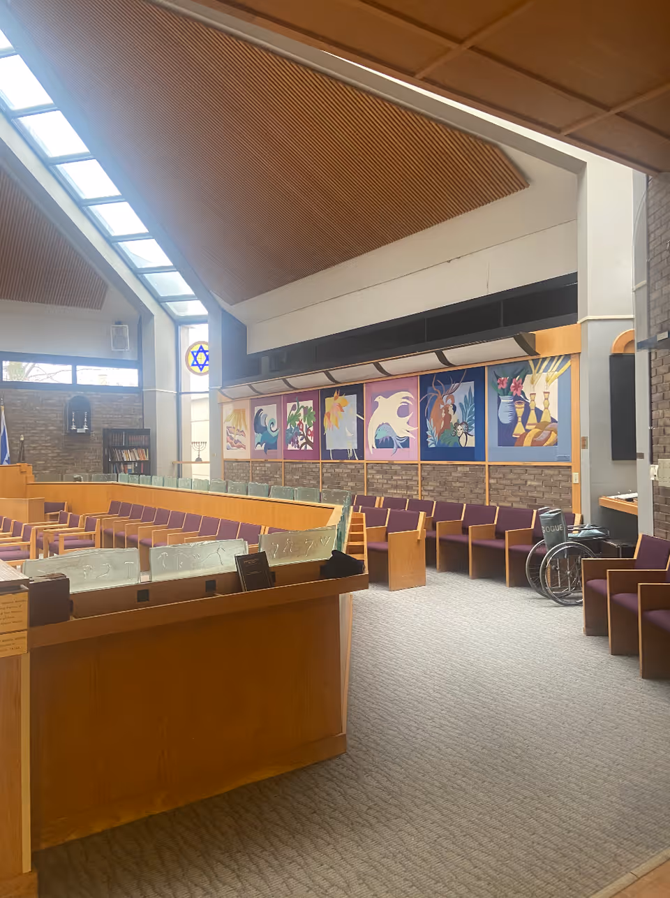 Interior of a chapel or worship space with wooden pews featuring purple cushions, a slanted ceiling with skylights, colorful religious artwork on the wall, and a wheelchair near the seating area.