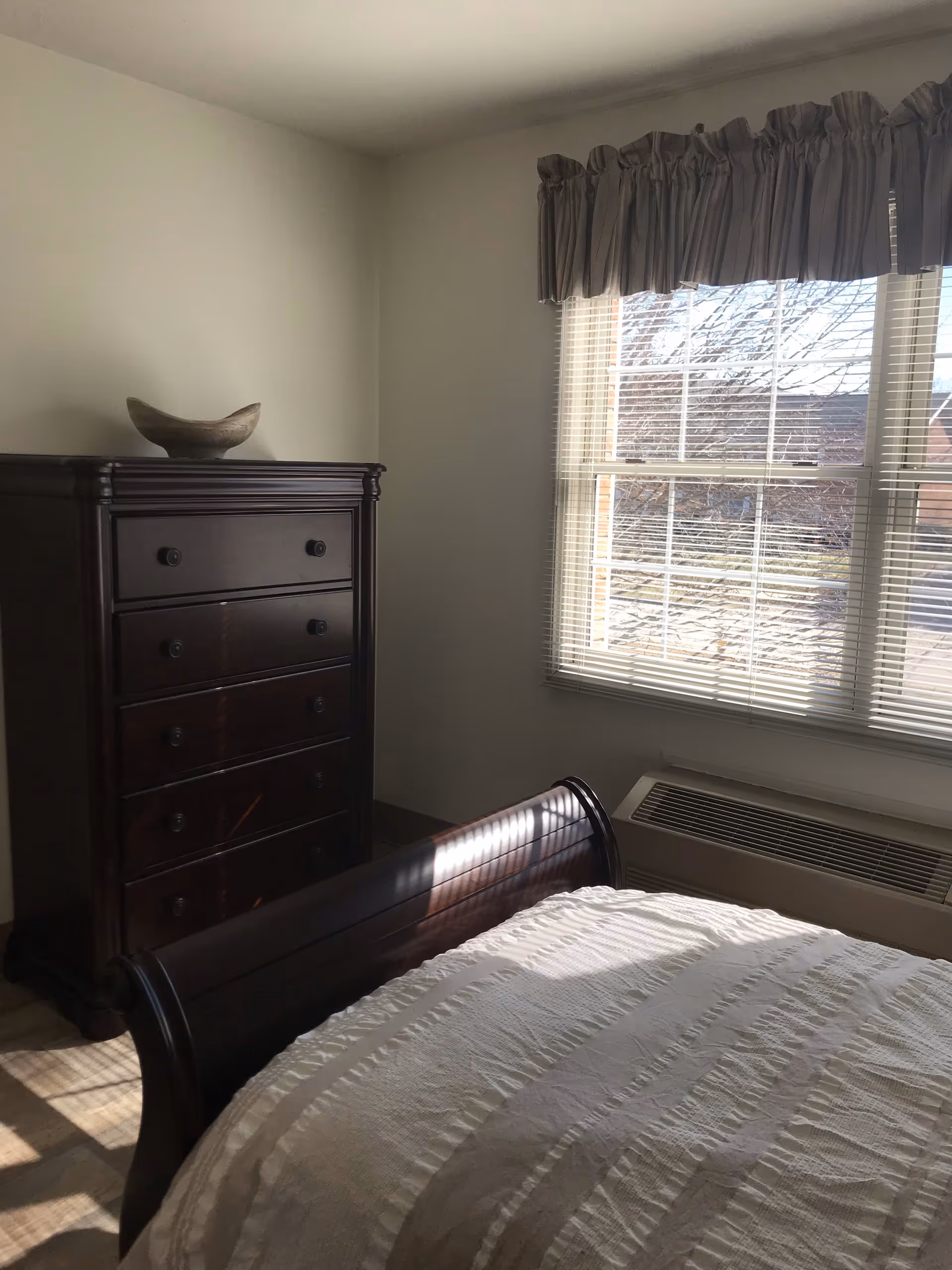 A bedroom with a wooden sleigh bed covered in a white textured bedspread, a dark wooden chest of drawers with a decorative bowl on top, and a window with blinds and a ruffled valance letting in natural light.
