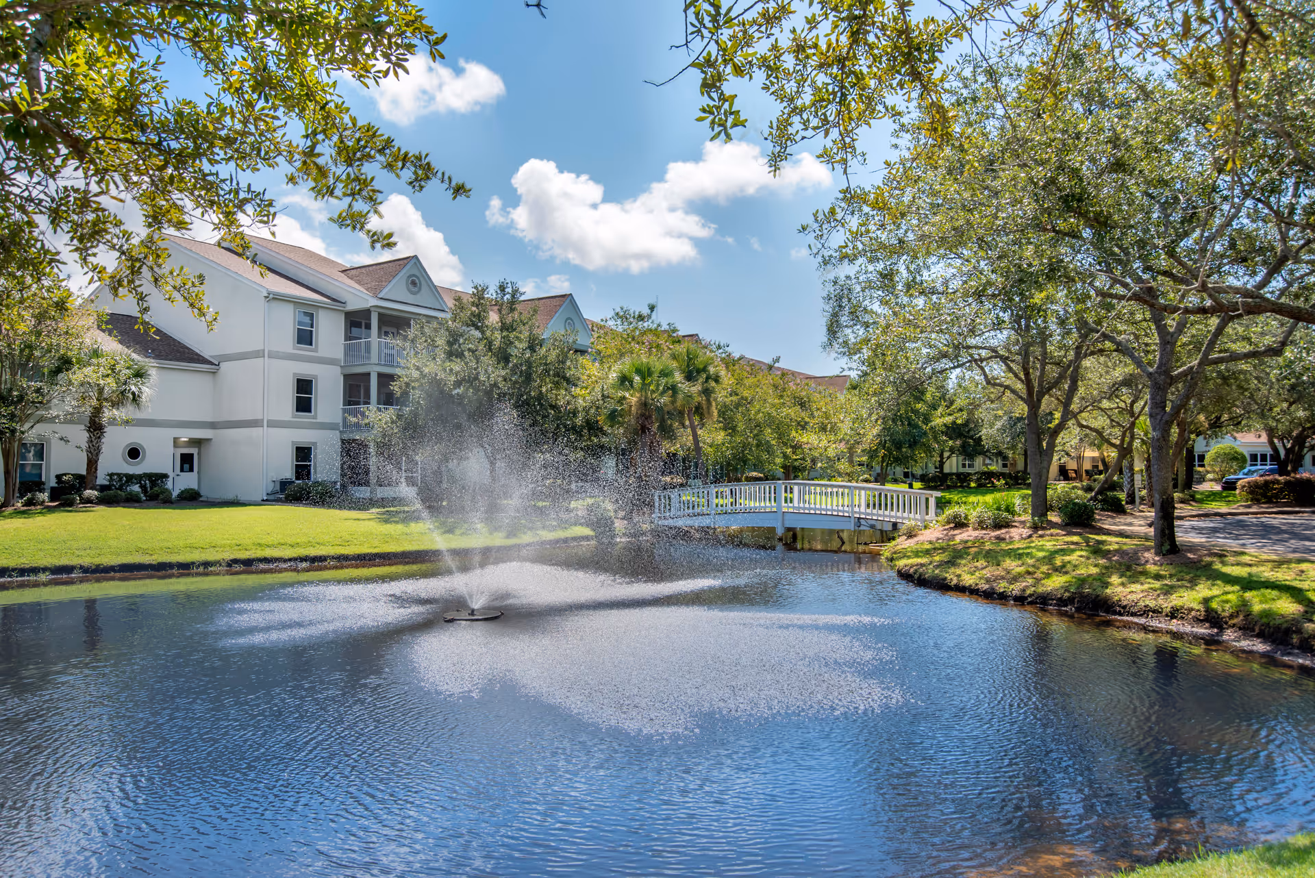 A serene outdoor scene at Brookdale Destin featuring a small pond with a water fountain spraying water upwards. A white wooden footbridge crosses over the pond, surrounded by green grass, trees, and shrubs. In the background, there is a multi-story building with balconies under a blue sky with scattered clouds.
