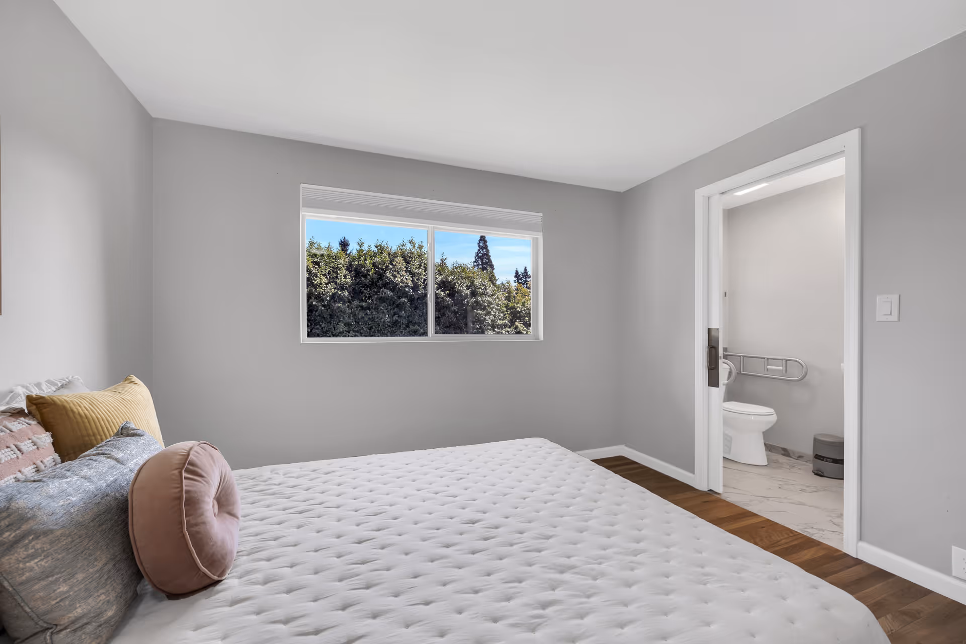 A bedroom with a large bed covered in a white quilt and several pillows in shades of pink, yellow, and gray. There is a window showing green trees outside. An open door reveals an adjacent bathroom with a toilet and grab bars.